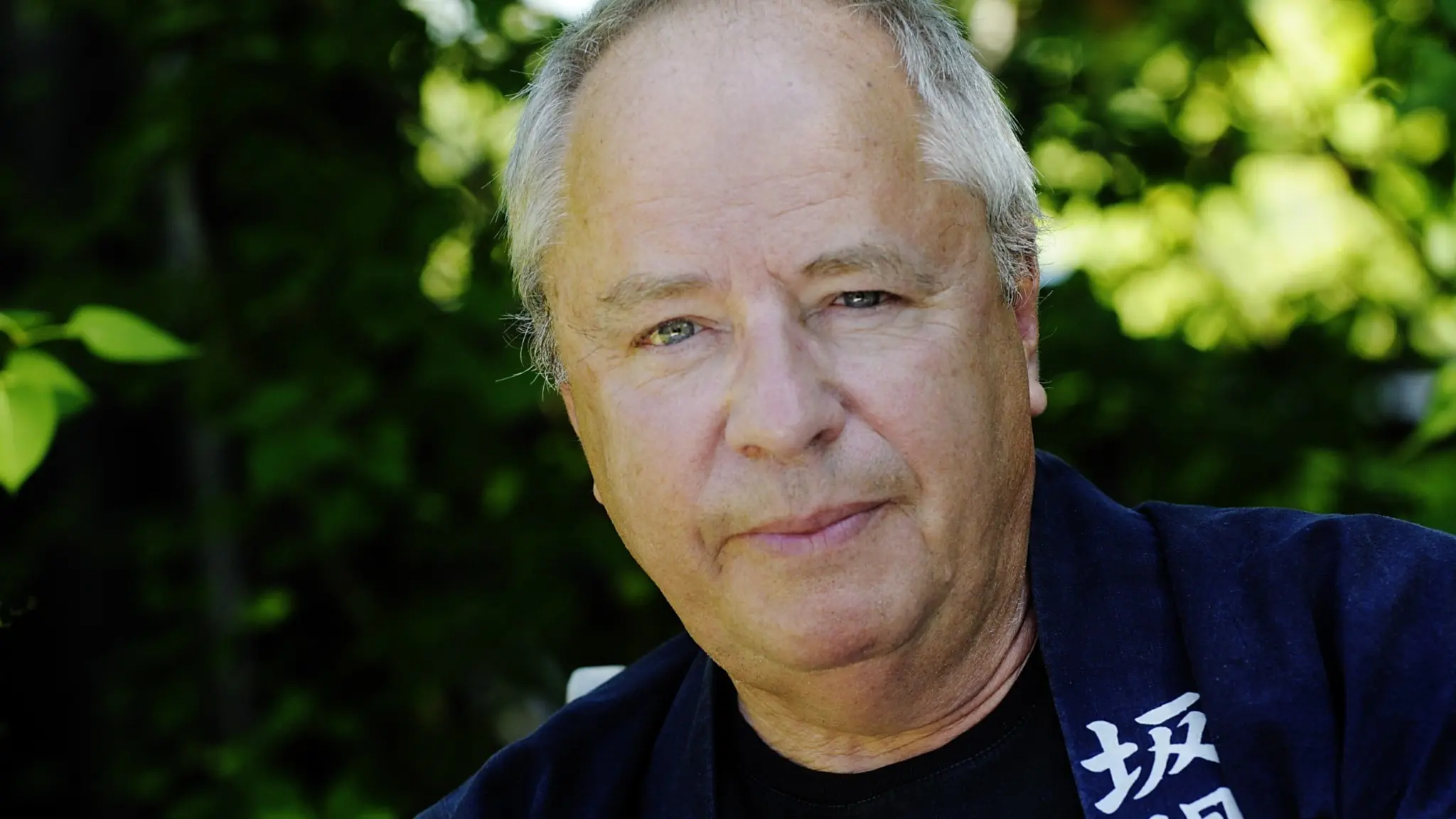 En person klädd i mörka kläder står utomhus framför en grönskande bakgrund. Ljuset skapar kontraster mellan skuggor och solbelysta områden i miljön.