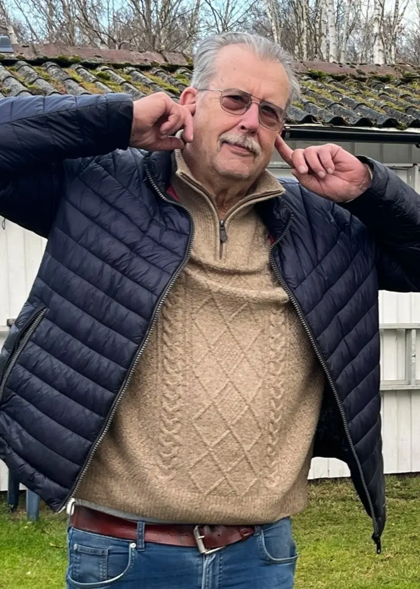 Lars Franzén från Mellbystrand står utomhus i en mörk jacka vid en byggnad, medan en röd lastbil syns körande på E6 i bakgrunden. Lars håller för öronen.