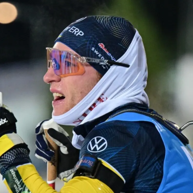 Två manliga skidskyttar i tävlingsdräkter med gevär på ryggen står på en belyst arena under en tävling. De interagerar med varandra, och sponsorskyltar syns i bakgrunden.
