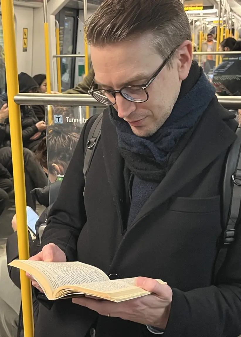 Erik, en man i svart kappa och ryggsäck, står och läser en bok på en tunnelbana. Bakom honom syns passagerare som sitter och använder sina mobiltelefoner.