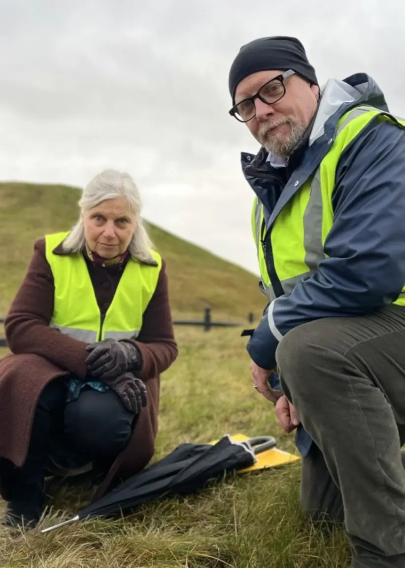 Två personer i reflexvästar undersöker en gräsklädd kulle, troligen en fornlämning. En av dem sitter på huk med utrustning på marken. Intill visas en närbild på gräs och jord.