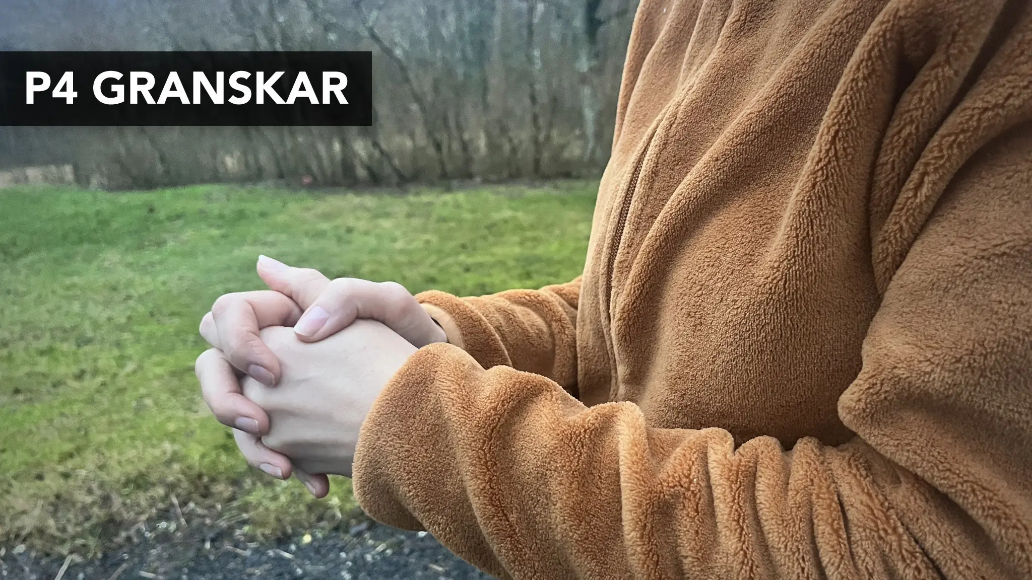 En person i brun fleecejacka står utomhus och håller sina händer samman, med en bakgrund av gräs, buskar och träd. Bilden kopplas till ”Sofia” och en förskoleskandal.