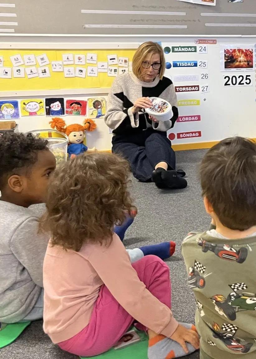 Pedagogen Elena Evertsson på förskolan Författaren i Vivalla sitter på golvet med en grupp barn och håller i en bok. Barnen deltar i en språkutvecklande aktivitet och omges av färgglada väggdekorationer.