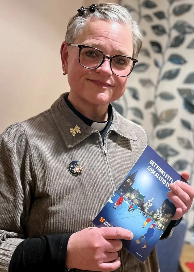 Tillförordnad kyrkoherde Brita Wennsten håller i ett julkort, ståendes framför ett fönster dekorerat med stjärnor och ljus, iförd en stickad tröja.