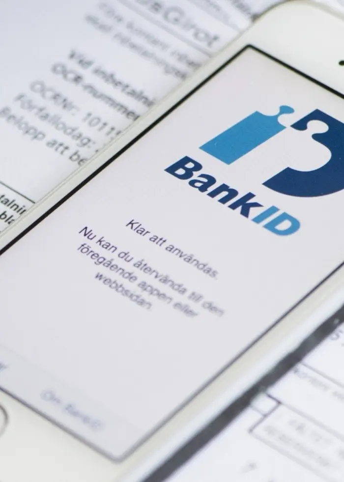 En smartphone med Bank-ID-appen visas på skärmen, placerad ovanpå pappersdokument med betalningsinformation. Bilden symboliserar digital hantering av ekonomiska ärenden.