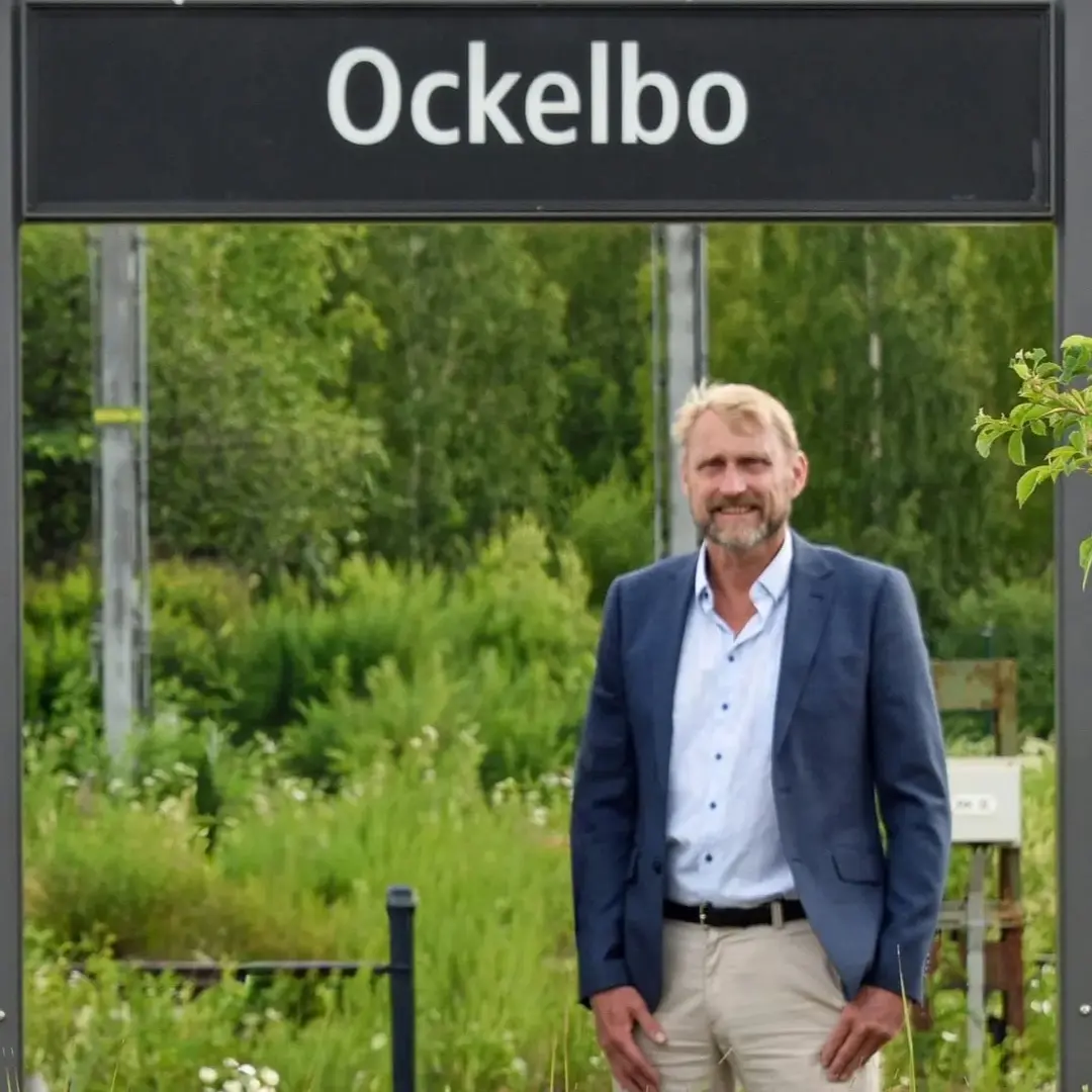 Ockelbos kommunchef Joachim Krüger står vid en skylt med texten 'Ockelbo' på en grönskande plats med en röd byggnad i bakgrunden, klädd i ljusa byxor och blå kavaj.