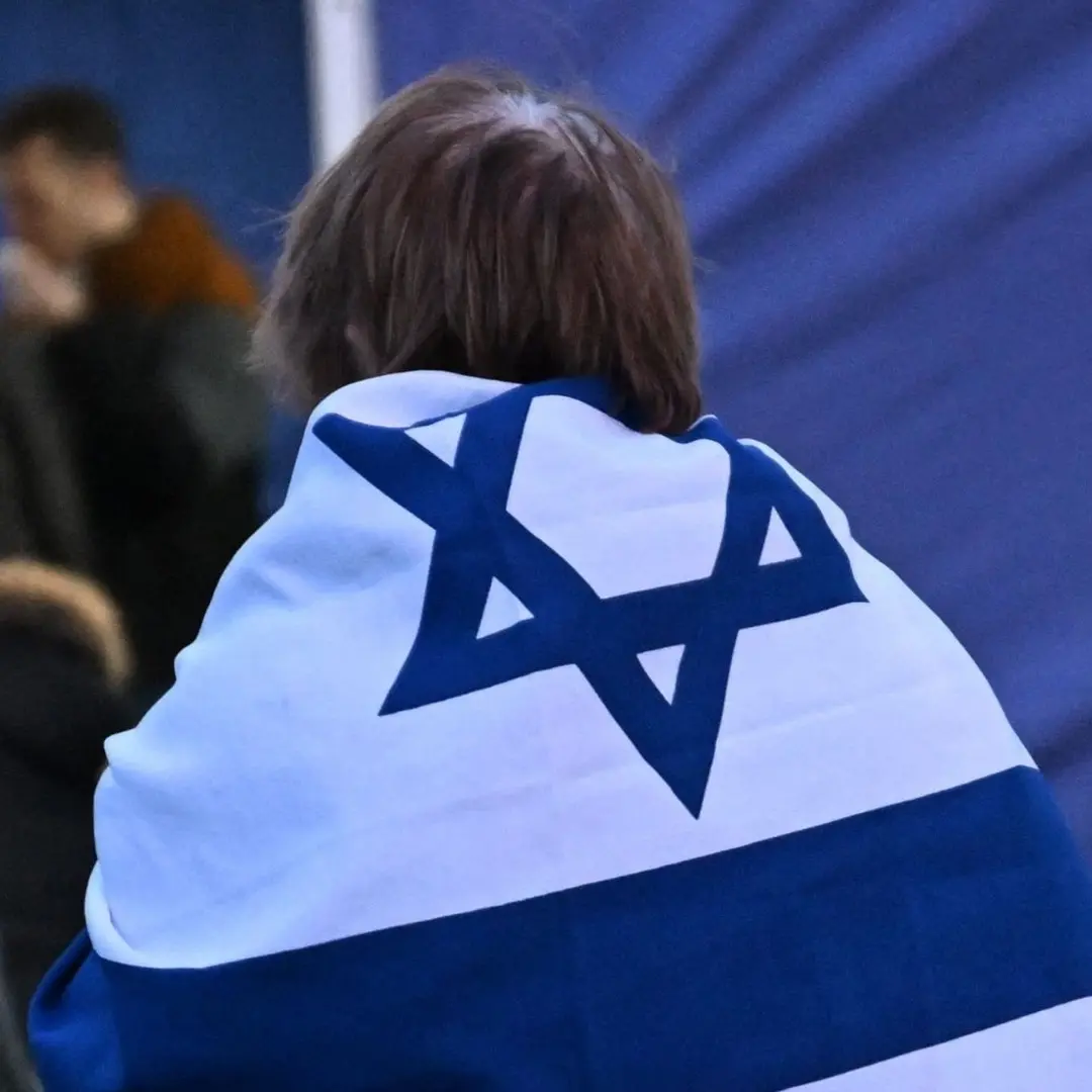 En person med en israelisk flagga över axlarna står i en folksamling. Människor och tältstrukturer syns i bakgrunden. Scenen verkar vara utomhus och har en allvarlig atmosfär.