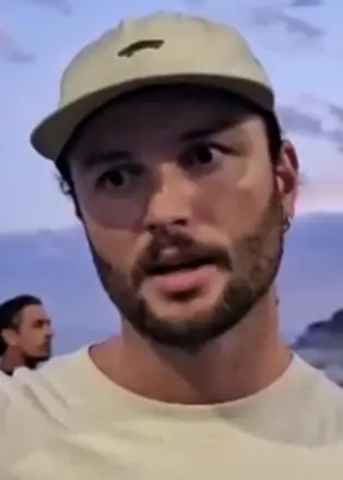 En person står utomhus vid havet under en solnedgång, klädd i en beige t-shirt med texten 'Arvust' och en keps. Personen håller upp något i handen. I bakgrunden syns gräs och en kustlinje.