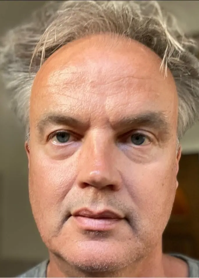 En person med grånat hår syns mot en suddig bakgrund inomhus. Bakgrunden består av neutrala färger och möbler som antyder ett hem.