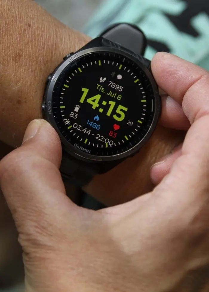 Närbild på en träningsklocka som visar tiden 14:15. En person justerar klockan medan hen sitter med gröna träningsskor synliga i bakgrunden.