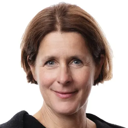 En person i skidkläder och en gul mössa står i en vintermiljö med skidstavar synliga i bakgrunden. Bredvid står en person i mörka kläder med armarna i kors. Snöfall kan ses i bakgrunden.