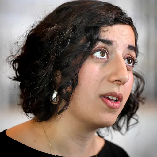 En person med lockigt hår står framför en blå bakgrund med ett svagt synligt emblem. Personen är klädd i en mörk tröja och håller en mikrofon i handen.