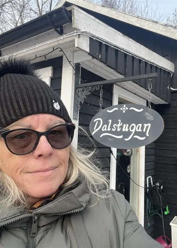 Ett snötäckt café med svart träfasad och dekorationer utanför, inklusive bord och krukor. Till vänster syns en person klädd i vinterkläder framför en skylt med texten 'Dalstugan'.