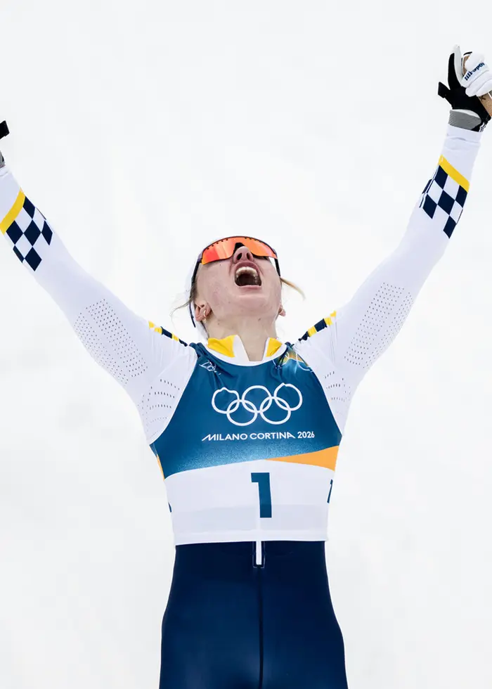 Tre skidåkare i tävlingsdräkter står med skidor och stavar i händerna, framför en bakgrund av snö och skog. De bär blå nummerlappar med olympiska ringar och ser ut att fira en framgång.