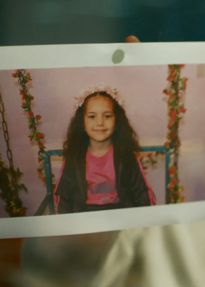 En person håller upp ett fotografi med en bild på en flicka med långt hår som sitter framför en rosa bakgrund dekorerad med blommor. I bakgrunden syns en suddig miljö med blåa och gula toner.