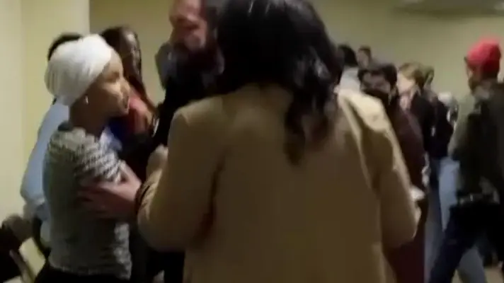 En person med vitblont hår och mörk kappa står utomhus med andedräkt synlig i den kalla luften. Bakgrunden är suddig med naturliga färger som antyder träd eller växtlighet.