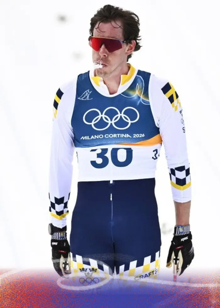 En längdskidåkare i svensk tävlingsdräkt har precis gått i mål efter femmilen vid vinter-OS i Val di Fiemme, Italien. Skidåkaren står på en snötäckt bana med markerade spår och blå koner i bakgrunden.