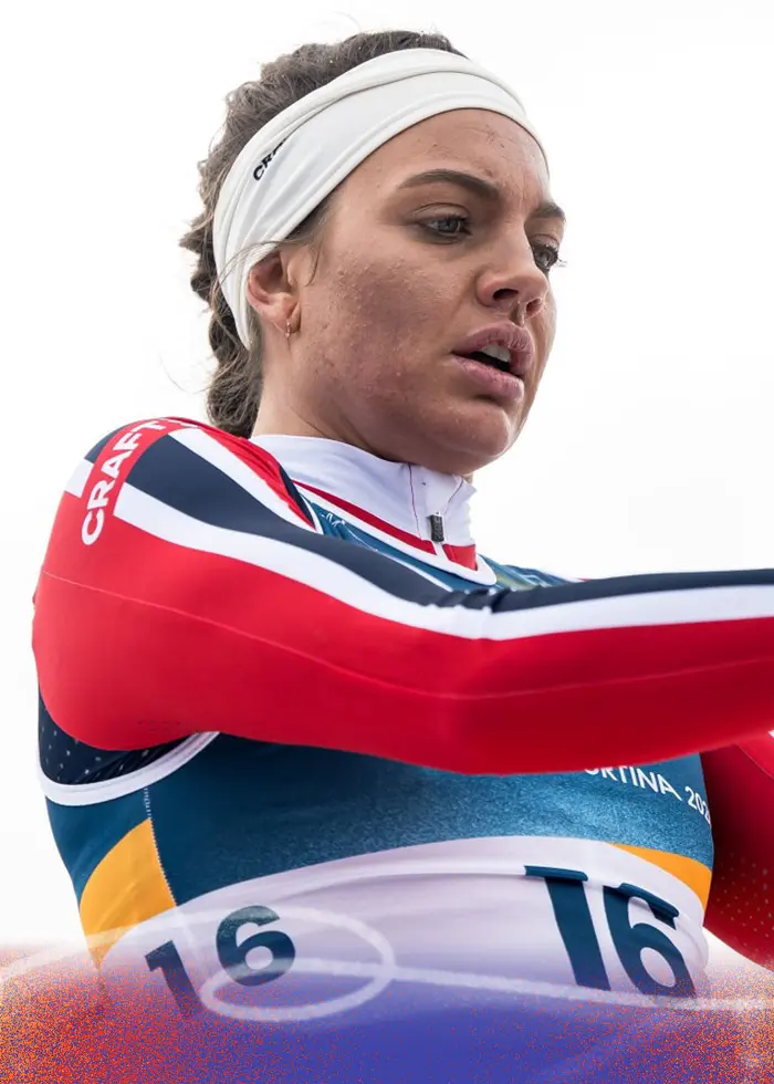 En längdskidåkare i röd och blå tävlingsdräkt syns i en aktiv position med stavar i händerna, omgiven av en ljus bakgrund. Nummerlappen visar siffran 16.