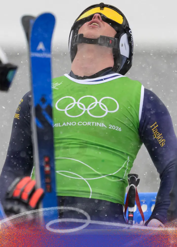 En alpin skidåkare i grön väst med olympiska ringar lutar huvudet bakåt i snöigt väder. Personen ser ut att uttrycka frustration eller besvikelse. Andra skidåkare syns i bakgrunden.