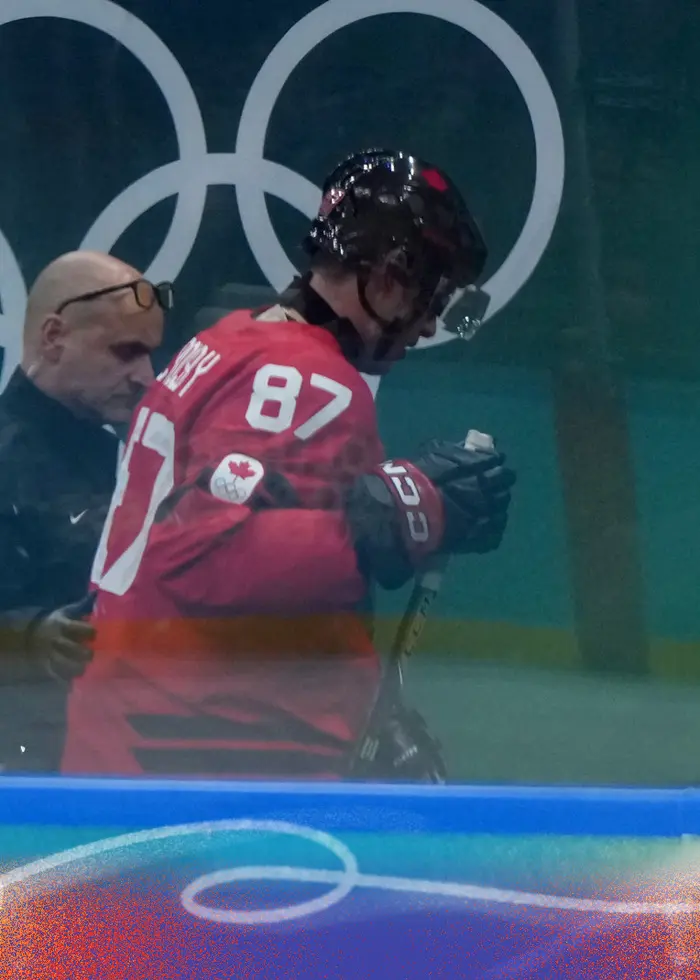 En ishockeyspelare i röd tröja med nummer 87 sitter på spelarbänken och ser ut att få vård. Till höger syns en person i kostym och slips i en ishockeyarena.