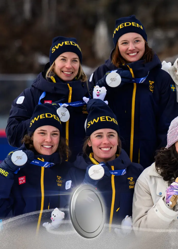 En skidskytt i svensk landslagsdräkt firar efter ett lopp med geväret på ryggen. Personen bär en blå tävlingsväst med olympiska ringar och nummer 34. Bakgrunden visar en skjutbana.