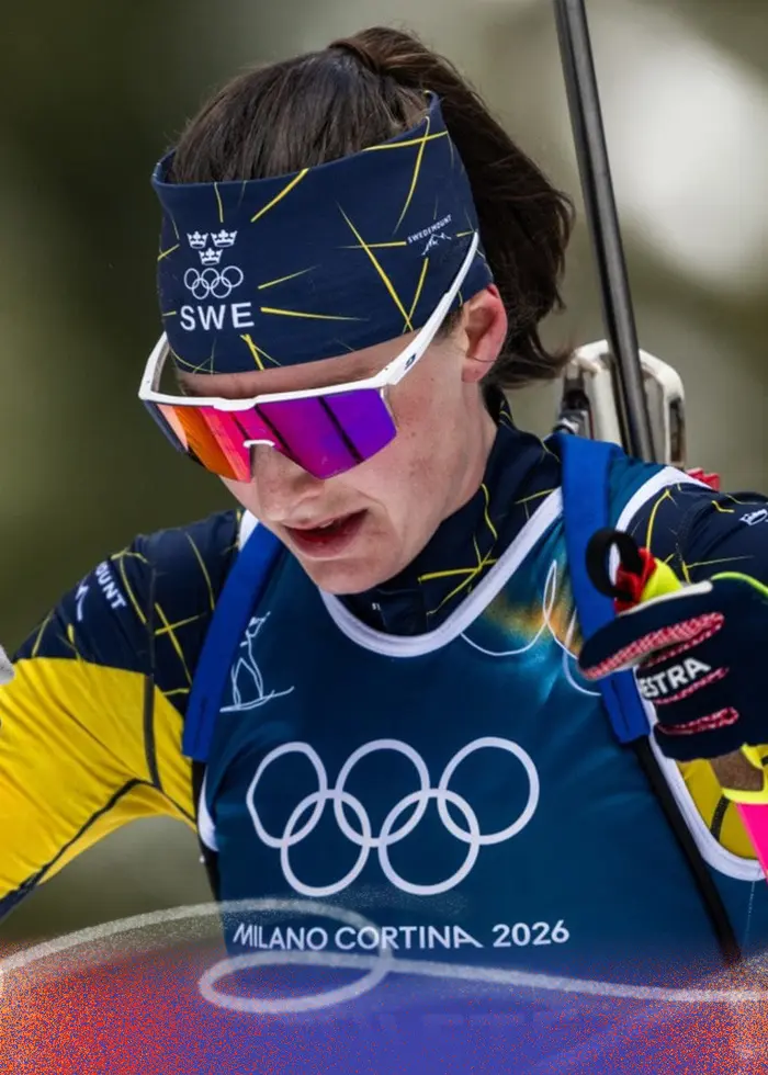 En kvinnlig skidskytt i färgglad tävlingsdräkt åker i en snötäckt skogsmiljö. Hon bär ett gevär på ryggen, har skidglasögon och koncentrerar sig på loppet. Olympiska ringar syns på hennes tröja.