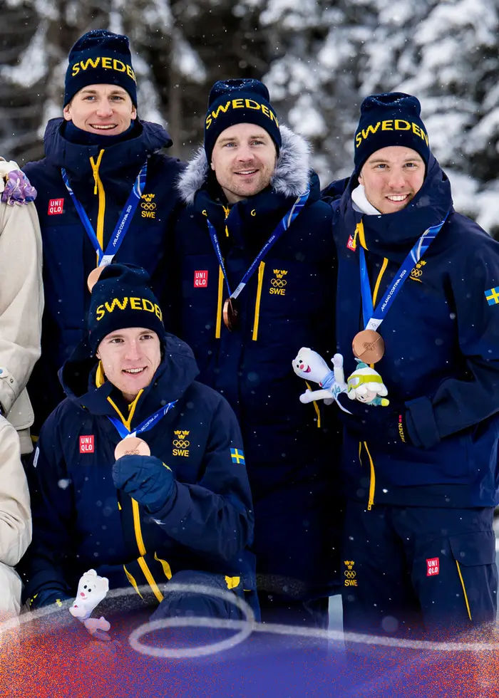 Fyra skidåkare i tävlingsdräkt håller sina skidor och poserar tillsammans framför kameran under ett snöigt vinter-OS. De bär nummerlappar och olympiska ringar syns på deras kläder.