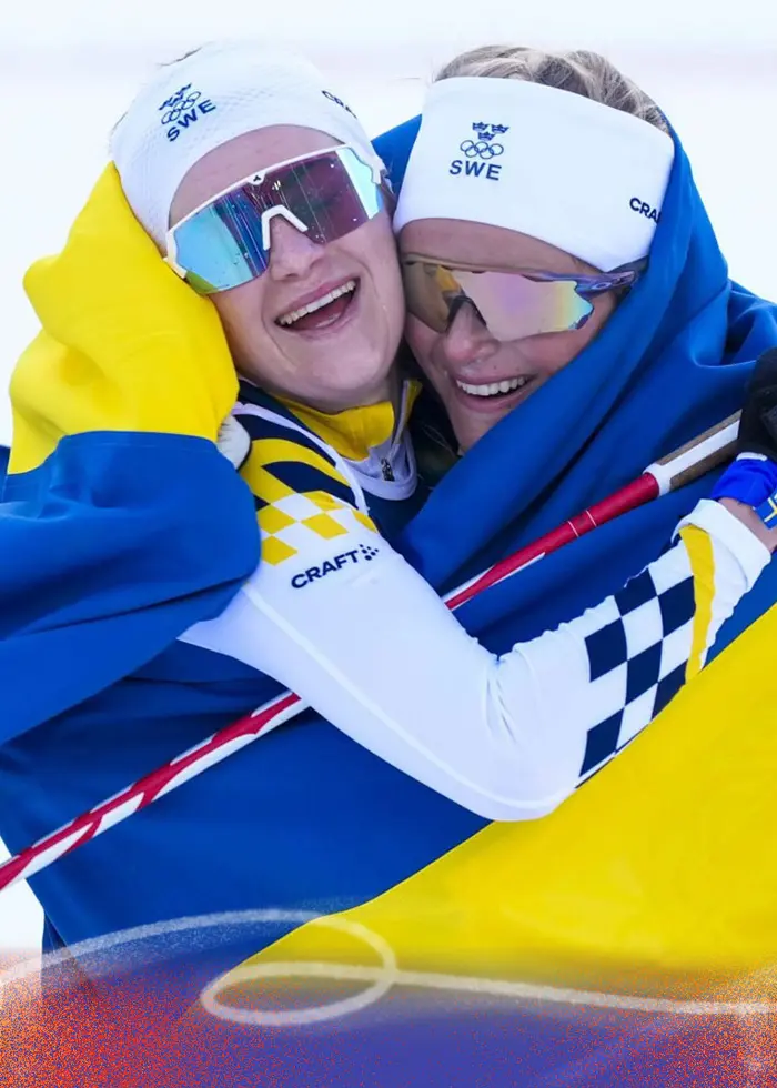 Två svenska längdskidåkare firar med den svenska flaggan efter en tävling vid Vinter-OS i Milano Cortina 2026. De håller om varandra på skidspåret och visar glädje över sin prestation.