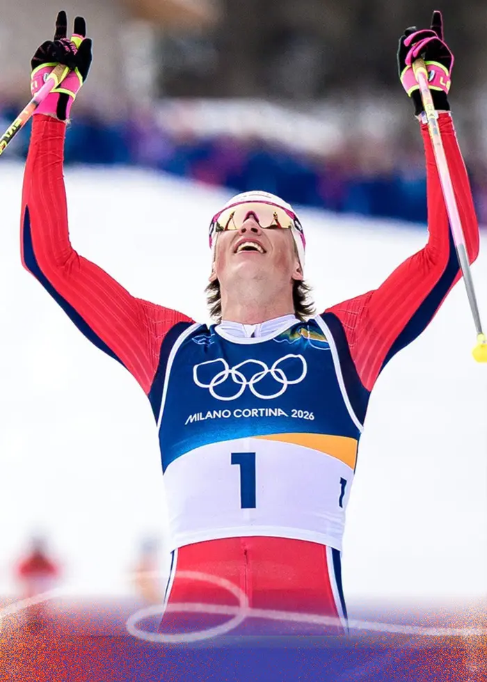 En längdskidåkare i röd dräkt och nummerlapp 1 sträcker upp armarna i segergest vid mållinjen under ett vinter-OS. Bakom syns en snötäckt bana och en publik i bakgrunden.
