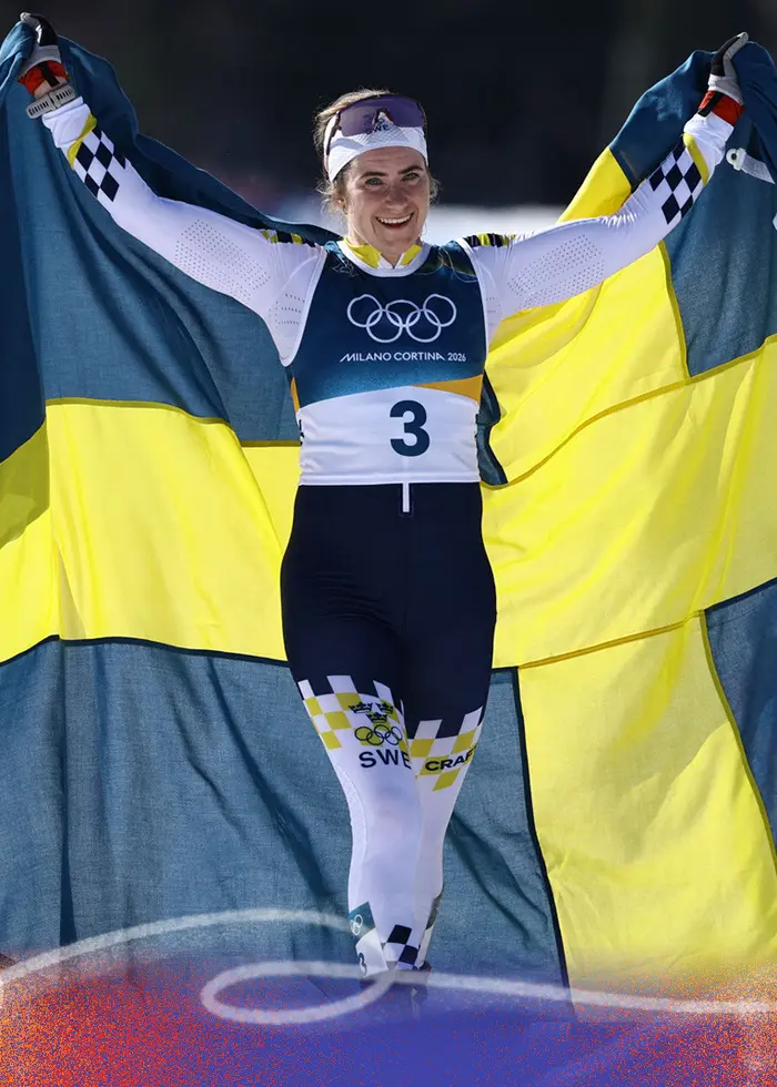 En skidåkare klädd i tävlingsdräkt firar med en stor svensk flagga över axlarna mot en bakgrund av snöklädda berg och blå himmel. Startnummer 3 syns tydligt på dräkten.