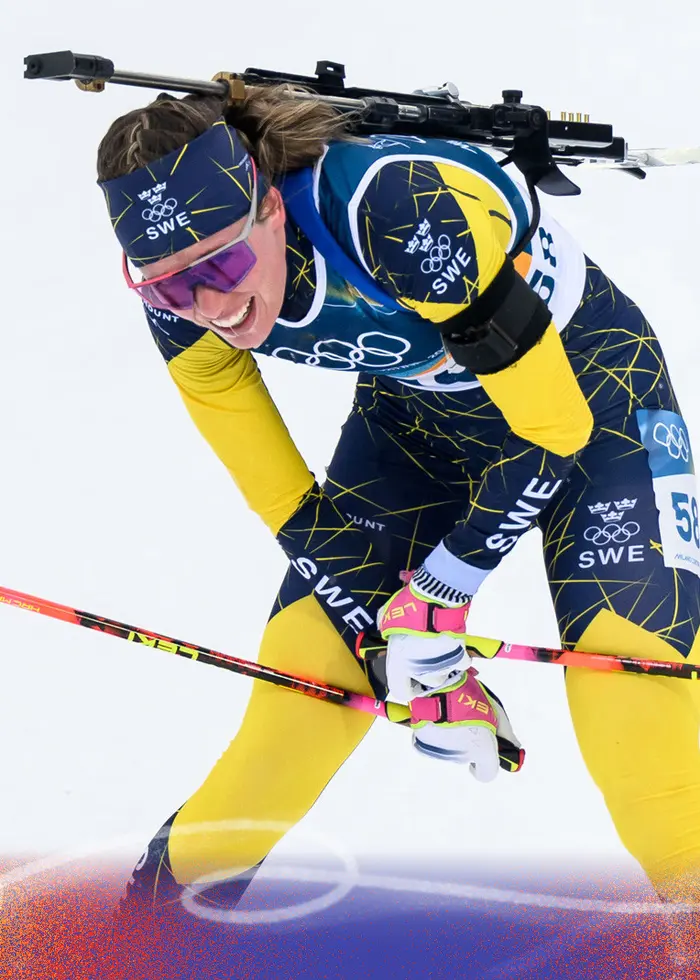 En kvinna i blå tävlingsdräkt och pannband med gula detaljer deltar i en skidskyttetävling. Hon bär ett gevär på ryggen och har sportglasögon på sig.