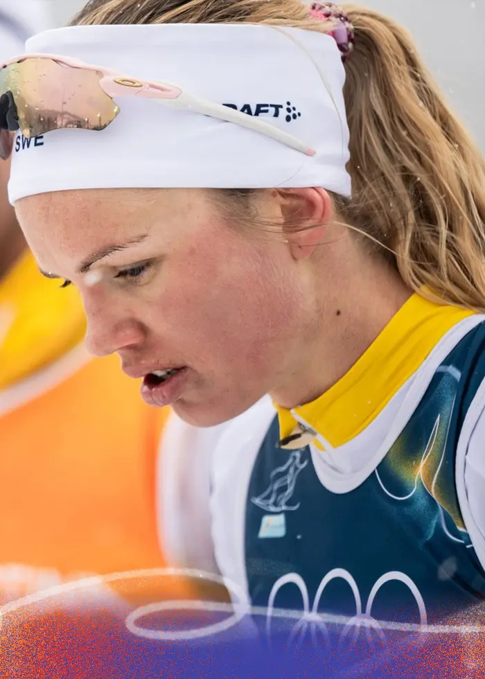 Två längdskidåkare i tävlingsdräkter står utomhus i vintermiljö. Den ena bär en gul väst och den andra en blå tävlingsdräkt med olympiska ringar. Snö faller lätt i luften.