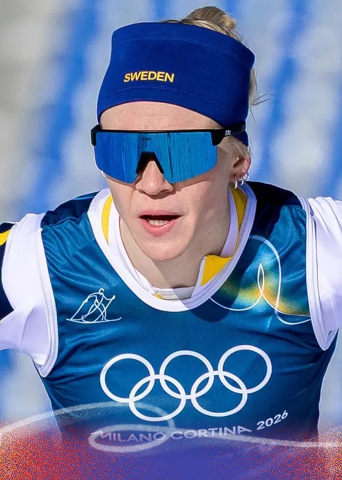 Två längdskidåkare i tävlingsdräkter och nummerlappar åker i ett snöklätt spår på en tävling. Bakgrunden visar en arena med tomma läktare.