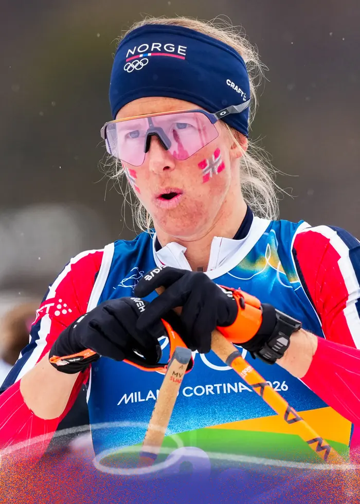En längdskidåkare i norsk landslagsdräkt ses kämpa i spåret under damernas 4 x 7,5 km stafett vid vinter-OS i Milano Cortina 2026. Hon bär blå pannband, rosa glasögon och håller stavar i händerna.