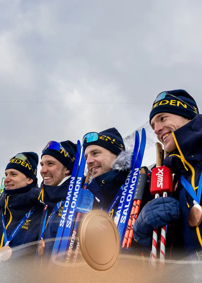 Fyra skidåkare i tävlingsdräkt håller sina skidor och poserar tillsammans framför kameran under ett snöigt vinter-OS. De bär nummerlappar och olympiska ringar syns på deras kläder.