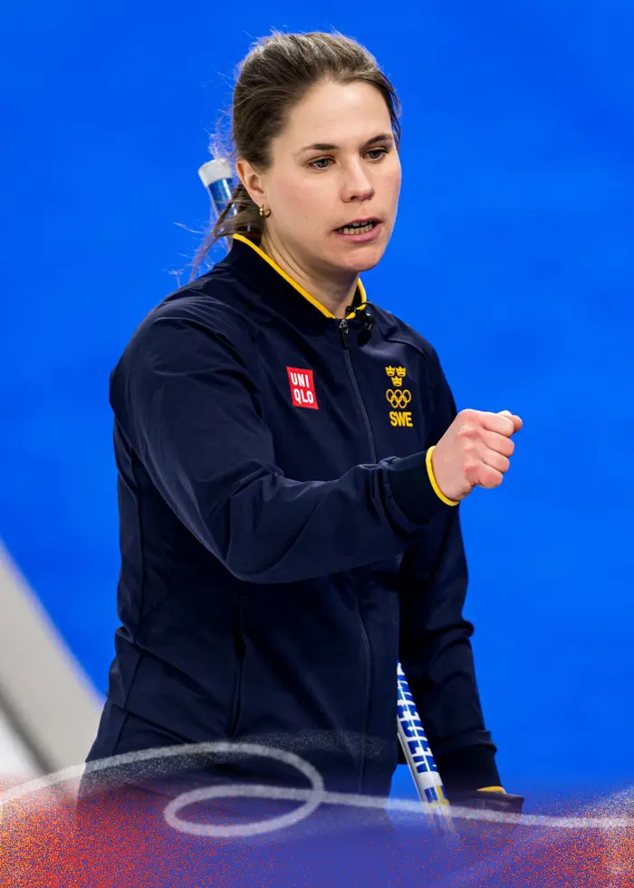 Bild från en curlingmatch med två svenska spelare i blåa tröjor som gör en gest mot varandra. I bakgrunden syns publiken och en person i vit jacka med röda detaljer.