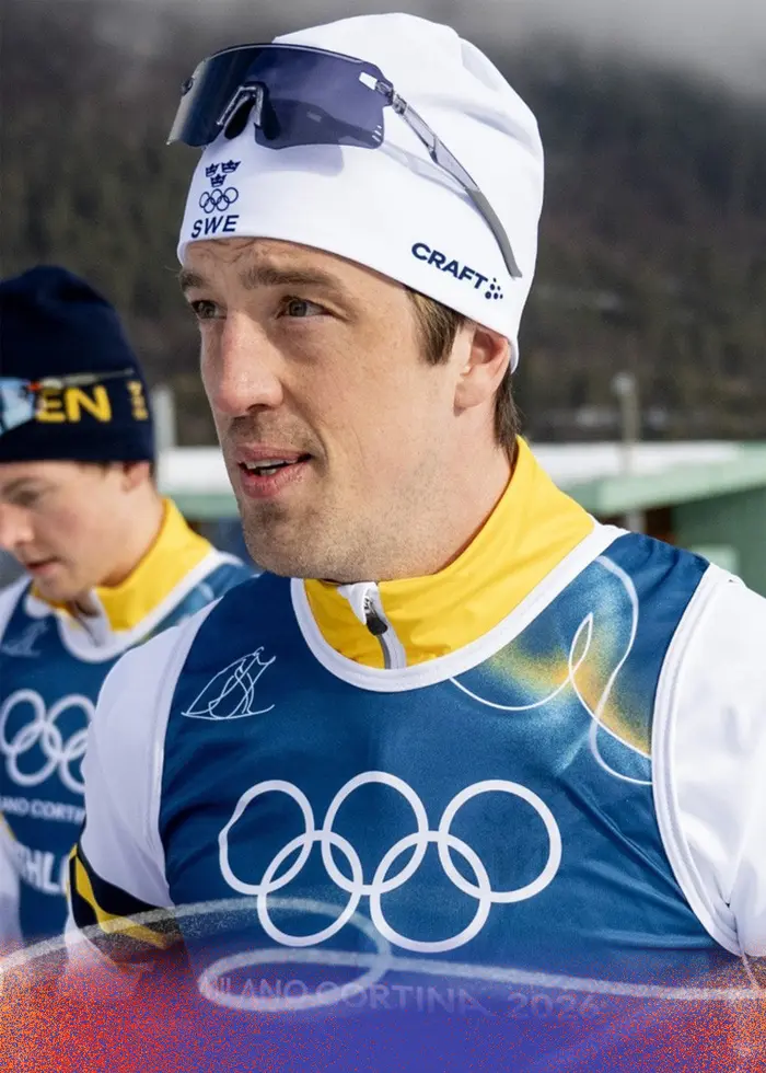 En skidåkare i vit dräkt och blå nummerlapp med texten 'ATHLETE' bär skidor på axeln under träning inför OS 2026 i Val di Fiemme, Italien. Snö och berg syns i bakgrunden.