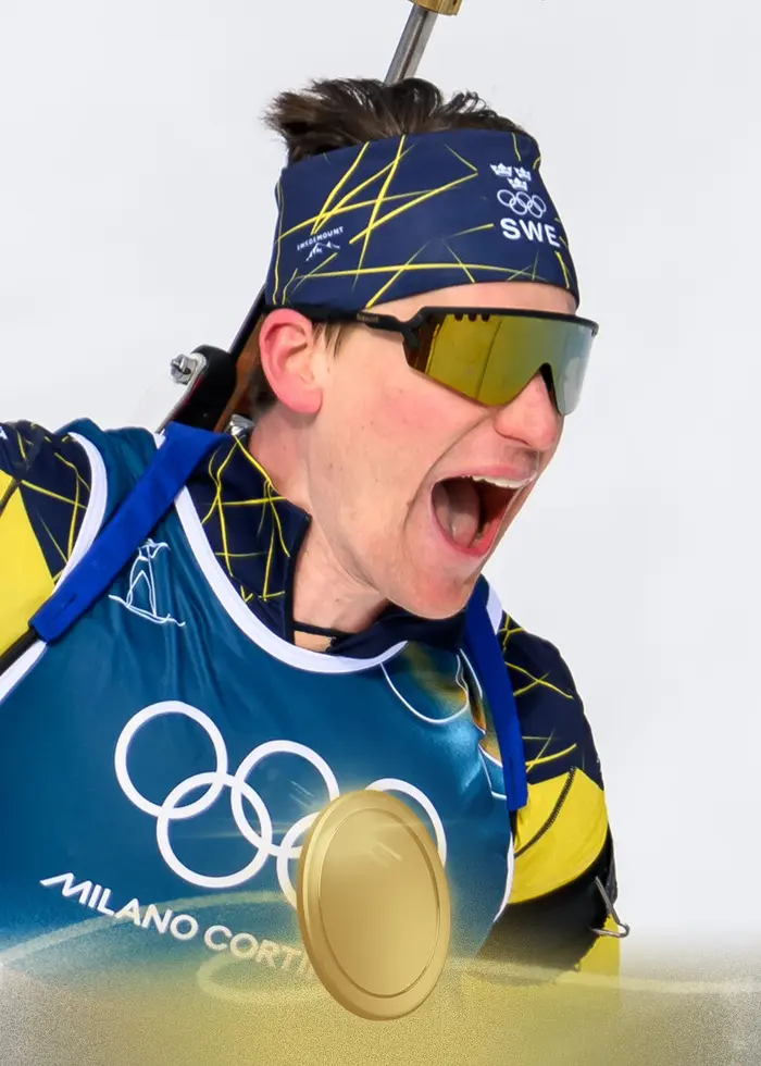 En svensk skidskytt i blågul dräkt firar guldet under vinter-OS 2026 i Anterselva efter herrarnas 12,5 kilometer jaktstart. Han håller en stav i ena handen och bär nummer 7.