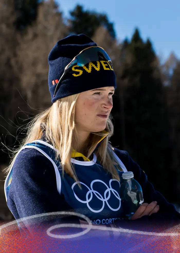 En längdskidåkare i gul och blå tävlingsdräkt håller stavar och har på sig vita handskar med Swix-logga. Personen deltar i vinter-OS i Milano Cortina 2026.