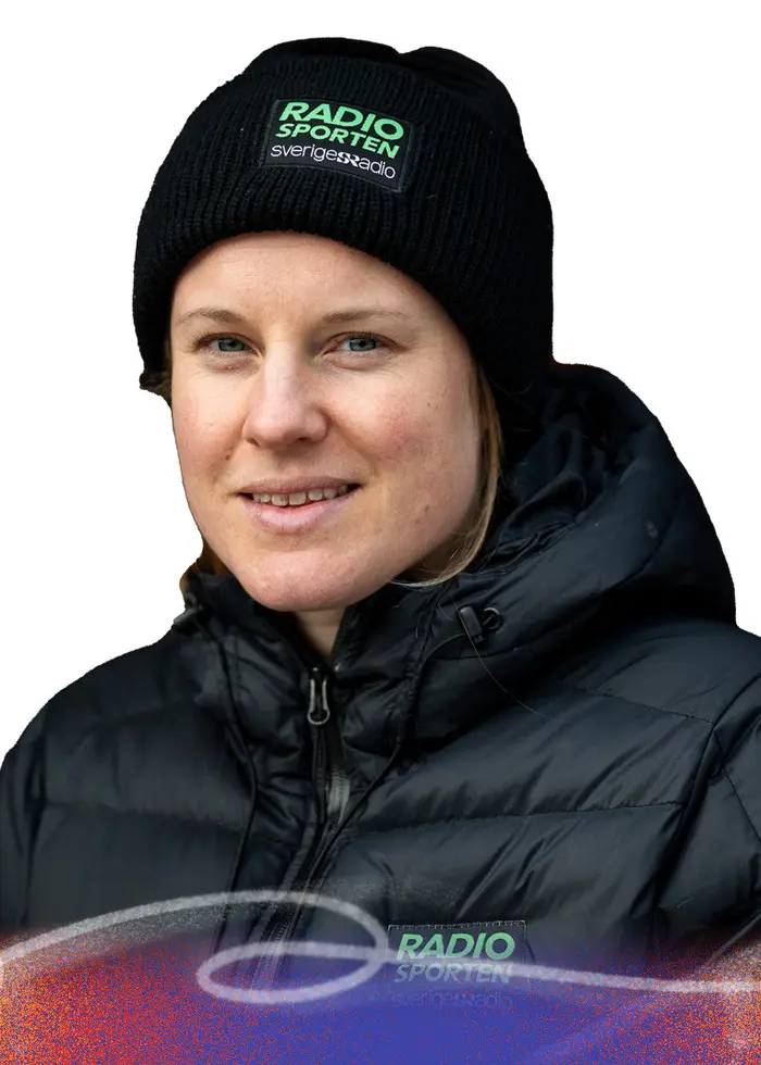 En längdskidåkare i tävlingsdräkt med svenska flaggan på ärmen och olympiska ringarna på bröstet åker skidor i snölandskap. I förgrunden syns en person i mörk jacka och mössa med text.