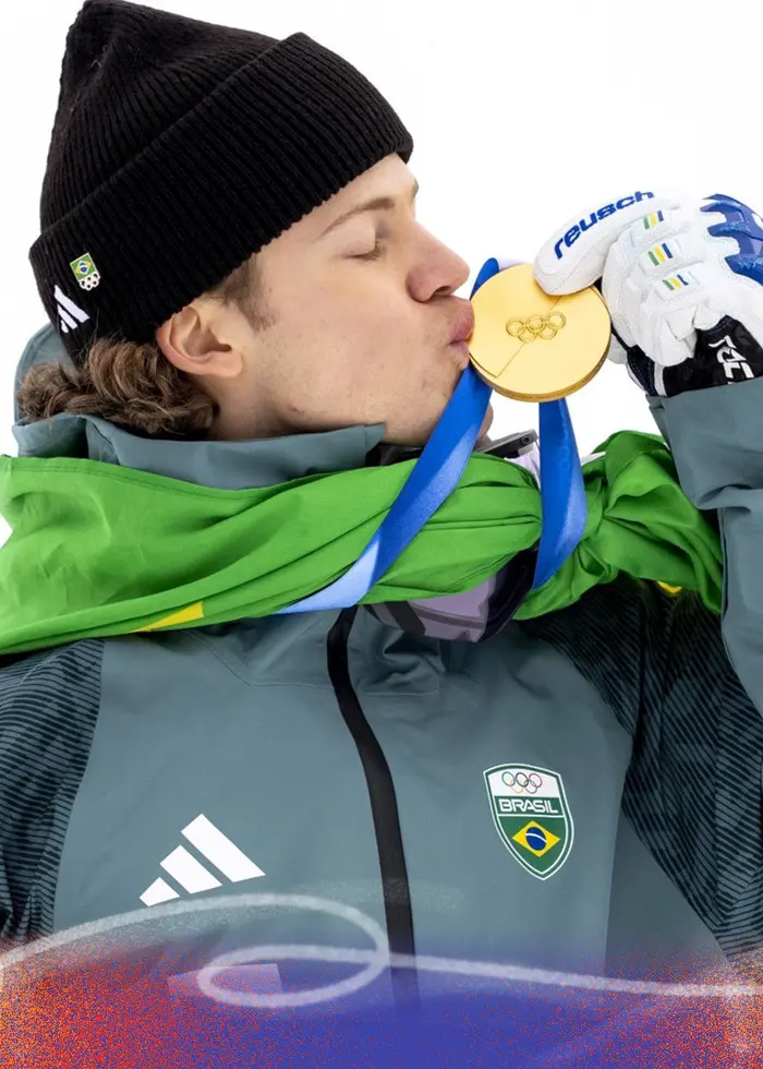 En alpin skidåkare iförd skidkläder kysser en guldmedalj efter en seger i storslalåm vid vinter-OS i Milano Cortina 2026. Personen håller en skida och är svept i en grön flagga.