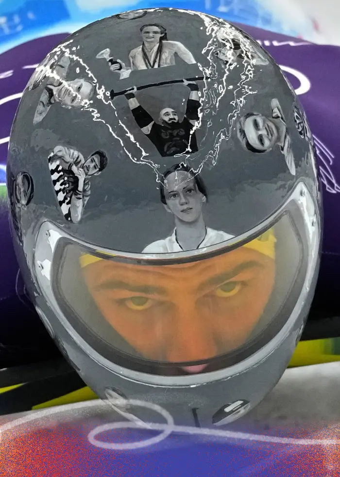 En skeletonåkare i lila och blå dräkt ligger på magen på en kälke under ett träningspass vid vinter-OS 2026 i Cortina d'Ampezzo, Italien. Hjälmen har en dekorativ design.