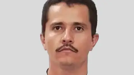 En person står vid en mikrofon med en knuten näve höjd i luften, iklädd en mörk topp. Bakgrunden är suddig och innehåller text eller logotyper.