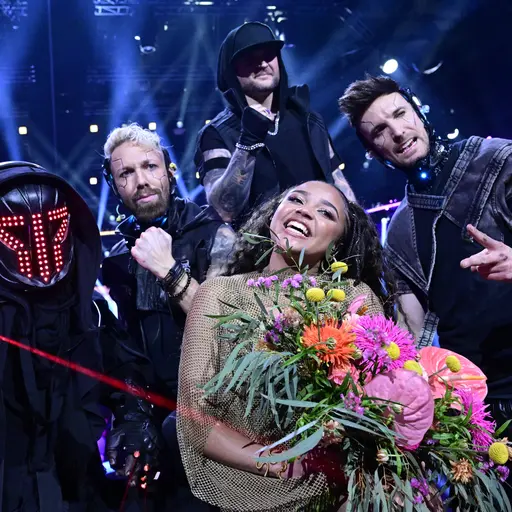 En grupp personer står på en scen under en färgstark ljusshow. En av dem håller en stor blombukett med färgglada blommor. Scenen är fylld med energiska poser och festlig stämning.