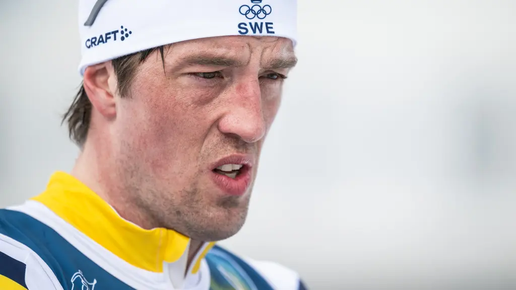 En manlig längdskidåkare i svensk tävlingsdräkt deltar i herrarnas stafett 4 x 7,5 km under vinter-OS i Val di Fiemme, Italien. Han håller skidstavar och bär startnummer 32.