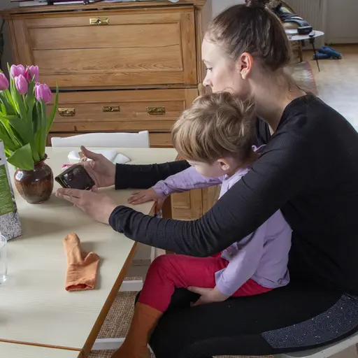 En kvinna med ett barn i knät sitter vid ett matbord och tittar på en telefon.