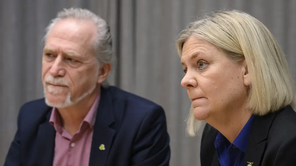 En person med ljust hår ses i närbild på en pressträff, klädd i en blå skjorta och kavaj. Bakgrunden är ljusblå och suddig, vilket ger fokus på personen i förgrunden.