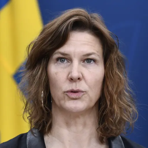 En kvinna står vid ett podium och talar under en pressträff. I bakgrunden syns en svensk flagga samt en EU-flagga.