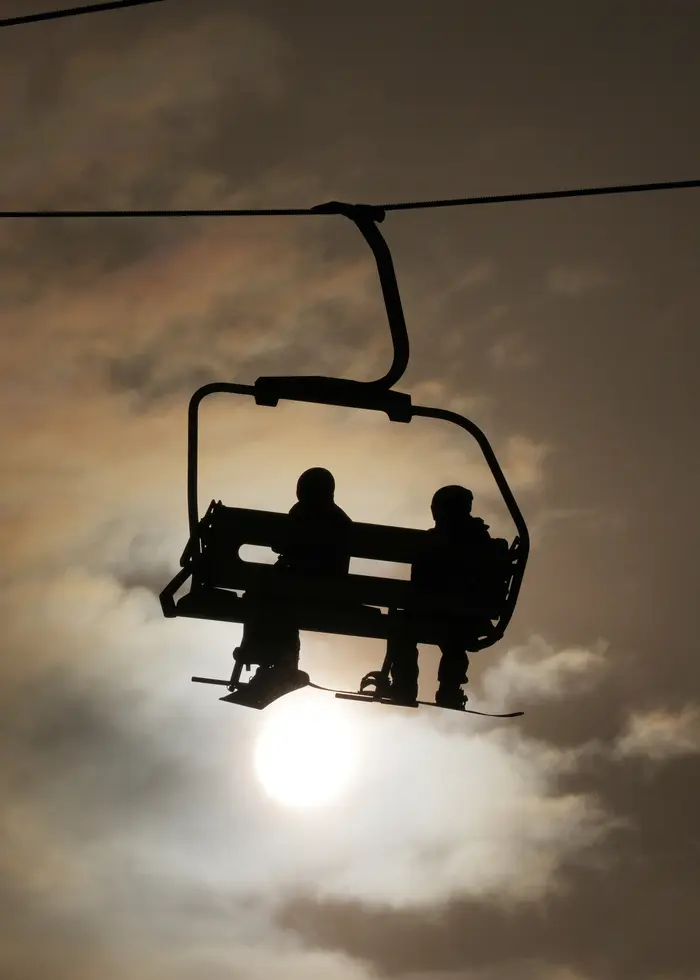 Två skidåkare är svarta siluetter i en stolslift för två personer. Himlen är mörk. Foto: 	Abbie Parr /AP/TT