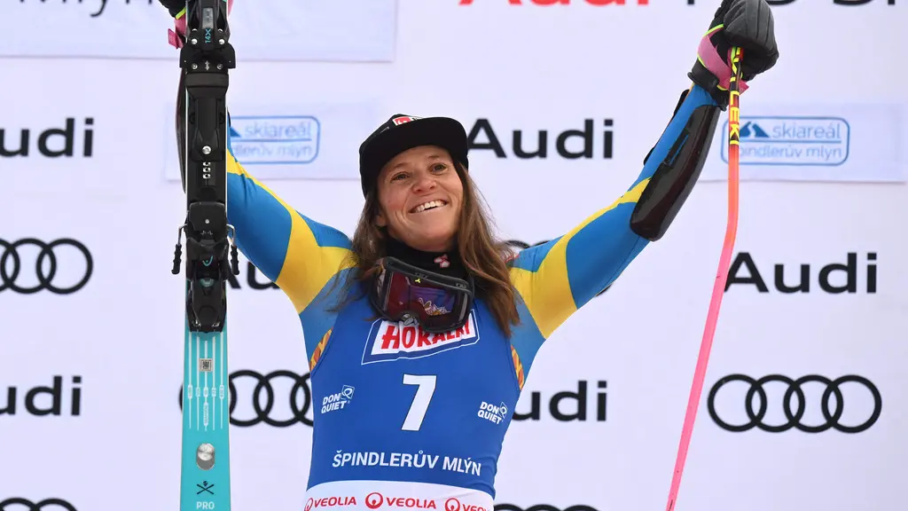 En alpin skidåkare i blå tävlingsdräkt svänger kraftfullt i en backe under en tävling. Till vänster syns en person i gul mössa och tävlingskläder med en medalj runt halsen.